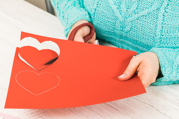 Valentine's day theme. Female hands cut the heart out of the paper. Packed gifts, tools on a battered wooden table. Workplace for the preparation of handmade ornaments.