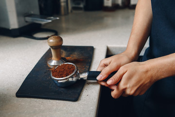 Young Caucasian woman barista using tamper to press ground coffee into portafilter to make espresso hot drink. Small local business work in cafe.