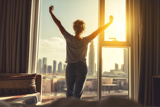 happy woman stretches and  opens curtains at window in morning