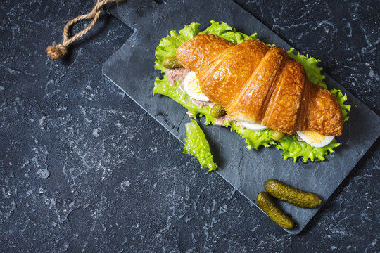 Croissant sandwich with tuna, hard boiled egg, salad and cucumber on stone table