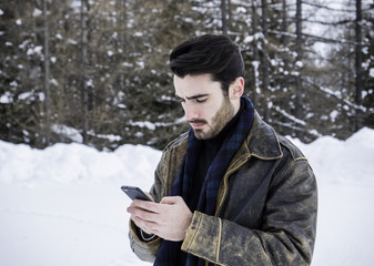 Handsome young man using smarpthone while posing among snowy woods in the mountain