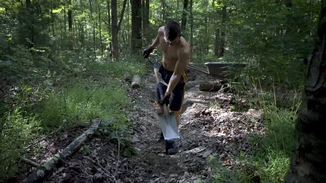 Young shirtless man digging dirt and rocks with a square shovel and putting it into a wheelbarrow.
