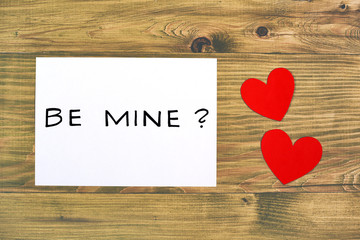 White paper with question be mine and heart shapes on wooden table.