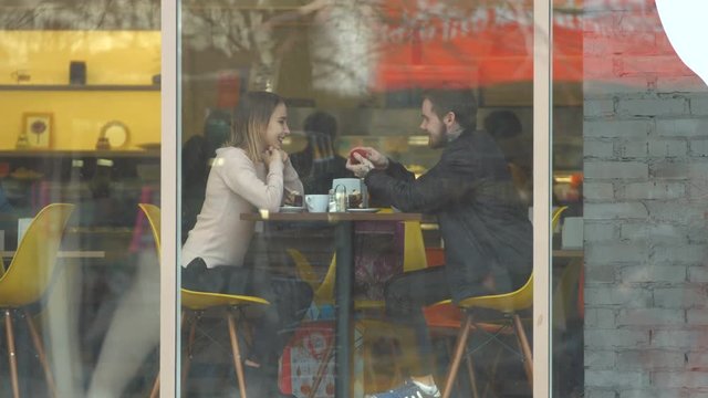 Young couple in a cafe.
Slow motion. A guy with a girl are sitting in a cafe behind the glass.
The guy gives his girl a small heart in which the ring.