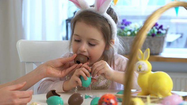 Little girl eating Chocolate Easter Eggs sitting at the holiday table with a basket and a yellow rabbit
