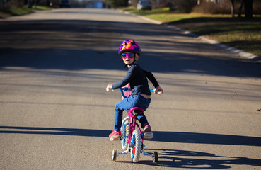A four year old girl in a helmet and fancy sunglasses pedalling her bicycle with training wheels down a street
