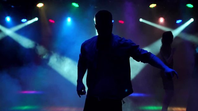 Silhouette of young guy dancing in a nightclub, in the background dancing young girls. Night party. A group of young people dancing and having fun in the dark on the background of flashing lights.