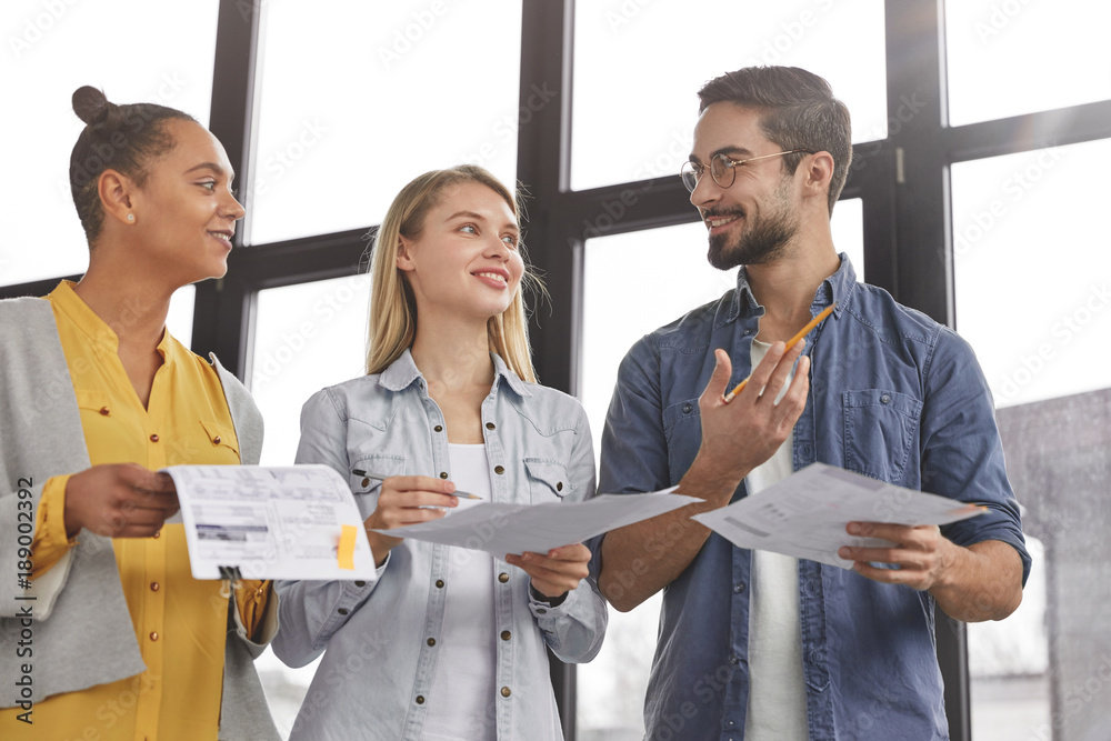 Wall mural group of friendly three multiethnic business people stand in office hall, hold papers, study financi