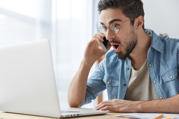 Portrait of stunned brunet male freelancer looks with stupefied expression into laptop computer, talks via smart phone, being pleasant astonished to recieve high money award, shares news with friend