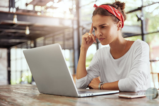 Beautiful female student focused into laptop computer, reads necessary information, prepares for examination, sits at wooden table of cafeteria. Thoughtful copywriter creats new company advertisment