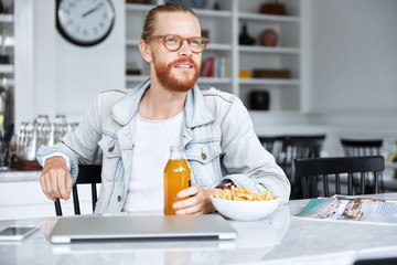 Thoughtful creative male designer wears spectacles, enjoys delicious lunch at kitchen, thinks about innovative ideas for new project, surrouded with modern electronic gadgets needed for work