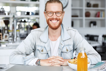 Pleased smiling hipster with ginger beard and mustache, wears spectacles, sits at table, has dinner break at cafeteria, being glad to finish work on laptop computer, expresses positive emotions