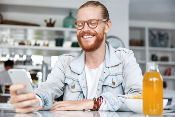 People, fashion, style and communication concept. Smiling pleased fashionable male wears spectacles, bracelet and denim shirt, glad to recieve message of congratulation from friend, has dinner at cafe