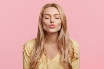 Lovely young woman with rounded lips and shut eyes, makes kiss, has long blonde hair, poses against...