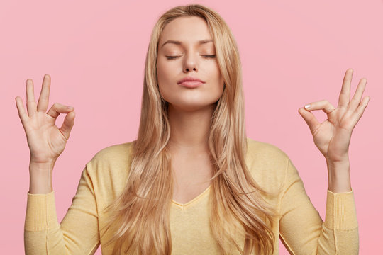 Blonde beauiful young female makes mudra sign, relaxes after hard working day, keeps eyes shut, practises yoga against pink background. Young pretty woman meditates indoor. Relaxation concept