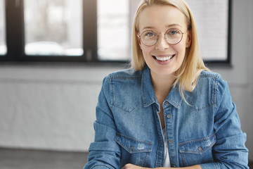 Indoor shot of smiling delightful blonde female copywriter being glad to finish her successful project work, looks gladfully at camera, wears spectacles, sits alone in cozy cabinet. Work concept