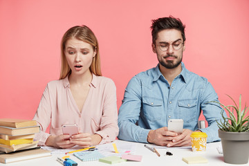 Pretty blonde woman with displeased look and handsome bearded male use modern electronic devices, connected to wireless internet, focused into screens. People, education, technology concept.
