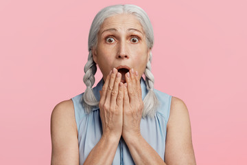 Frightened amazed grey haired female covers mouth, being shocked to hear bad news, expresses great surprsiment, scared of something, isolated over pink background. People and reaction concept
