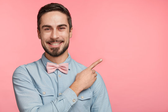Horizontal shot of pleased brunet male has appealing appearance, dressed in elegant shirt with pink bowtie, indicates aside with fore finger, has pleasant smile on face, isolated, copy area for text
