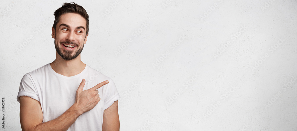 Wall mural Positive of brunet male with beard, has pleasant smile on face, dressed in casual white t shirt, isolated over concrete wall with copy space for your advertisment. Look at this! Stylish guy gestures