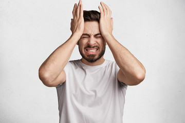 Young European bearded young male clenches teeth in desperation, regrets about something, dressed in casual t shirt, isolated over white background. Negative human emotions and feelings concept - obrazy, fototapety, plakaty