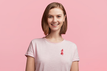 Fototapeta premium Healthcare and medicine concept. Smiling young famale with bobbed hairstyle wears pink breast cancer awareness ribbon, being involved in struggle against this serious disease, supports ill women