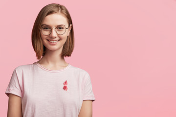 Cheerful female volunteer involved in helping people to cure disease, wears pink ribbon which symbolizes struggle from women`s disease mammary cancer, poses against pink background with copy space
