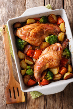 Delicious baked rabbit legs with potatoes, broccoli and tomatoes close-up in a baking dish. Vertical top view