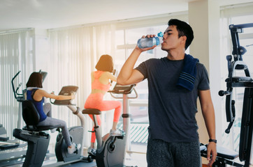 Fototapeta na wymiar muscular asian young handsome man with towel drinking water while resting after workout for good healthy in fitness gym, bodybuilder, healthy lifestyle, exercise and sport training concept