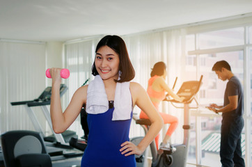 young asian pretty woman doing exercises with dumbbell at biceps in fitness gym with people background, bodybuilder, healthy lifestyle, exercise fitness, workout and sport training concept, soft focus