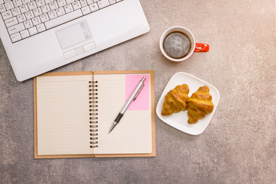 Laptop and notebook with coffee cup and croissant on the table