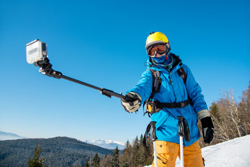 Male skier taking a selfie using his action camera and monopod in winter sunny day at ski resort