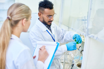 Portrait of two modern scientists working in medical laboratory doing research, focus on Middle-Eastern man pouring colored liquid to test tubes
