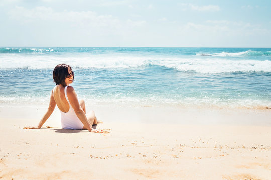 Woman in white swimsuit sits on empty ocean beach in sunny day