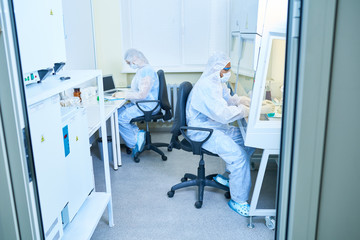 Full length side view of two scientists wearing protective suits and masks working at opposite tables in research laboratory
