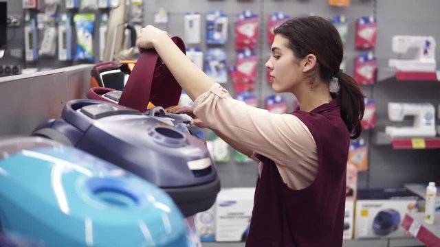 Girl choosing vacuum cleaner in appliance store. Coming up to a display row taking a dustbag out of vacuum cleaner. Costumer examining domestic equipment.