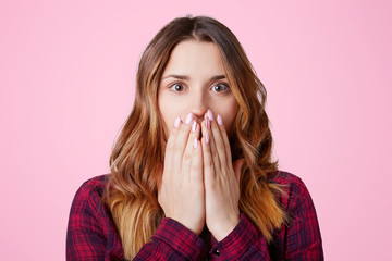 Amazed scared young pretty female dressed in checkered shirt, covers mouth with hands, being...