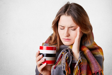 Frustrated ill young woman covered in warm blanket, keeps hand on temple, suffers from terrible headache, drinks hot beverage, tries to warm herself, has high temperature and flu, poses indoor