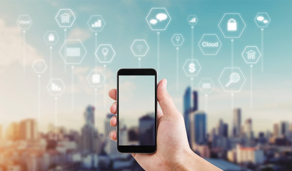 Hand using mobile smart phone with application icons such as online message, internet payment, banking, online shopping and etc. city sunrise background