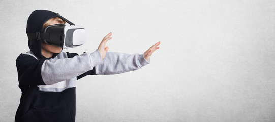 Little male child in sweatshirt and virtual reality glasses gestures with hands, stretches them forward, interacts with virtual environment, isolated over white concrete wall with copy space for text