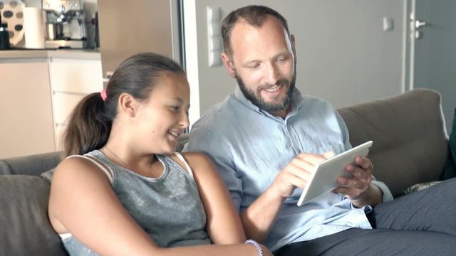 Father with his daughter talking and using tablet on sofa at home
