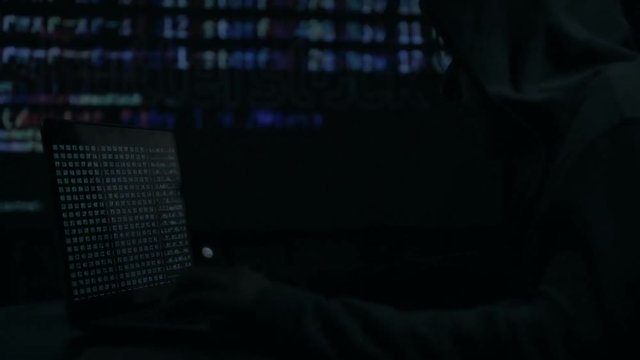 Male hacker working on a computer while green code characters reflect on his face in a dark office room.