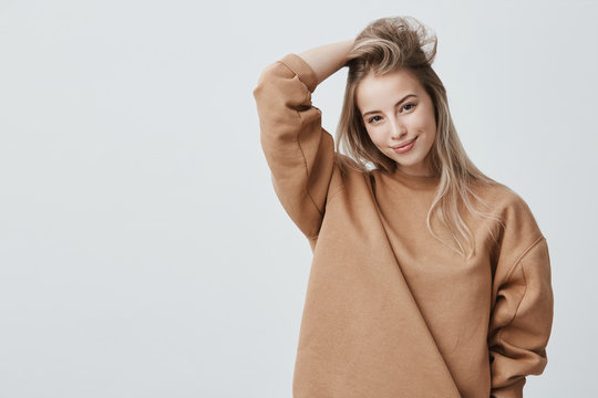 Young attractive woman wearing stylish long-sleeved sweatshirt looking with her dark appealing eyes at camera, smiling gently. Blonde caucasian female student posing against gray studio wall