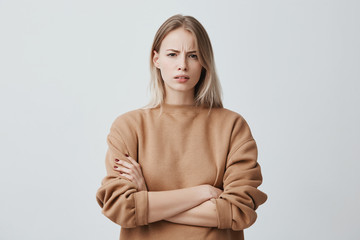 Waist-up portrait of beautiful girl with blonde straight hair frowning her face in displeasure, wearing loose long-sleeved sweater, keeping arms folded. Attractive young woman in closed posture. - 189033796