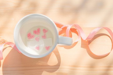 Paper cut of heart shape painted with red pencil and white paper in white coffee empty or blank cup on wood background with pink bow / ribbon under sunlight showing love relationship on Valentines day