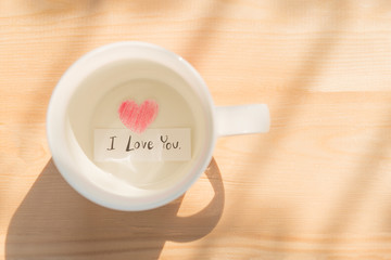 White coffee cup with paper cut in heart shape painted with red pencil and I love you in black on wood background under sunlight showing love relation concept on Valentines day or wedding celebration