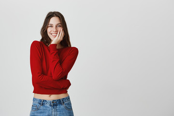 Close up of attractive young european woman with dark long hair in stylish casual outfit smiling touching face with hand, looking in camera with excited expression.