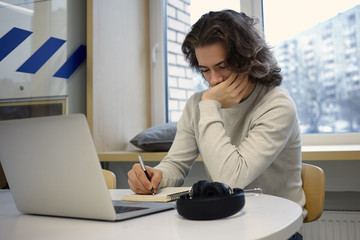 Unemployed young man dressed casually using laptop pc and making notes in his diary, writing...