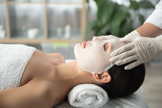 Side View Profile Of Tranquil Young Woman Lying On Table At Wellness Center. Beautician Is Applying Moisturizing Mask On Her Face