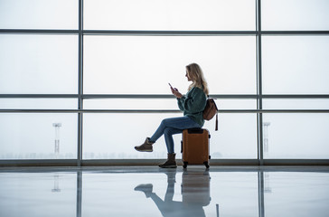 Side view of young woman waiting for the flight at the airport. She is sitting on suitcase and...